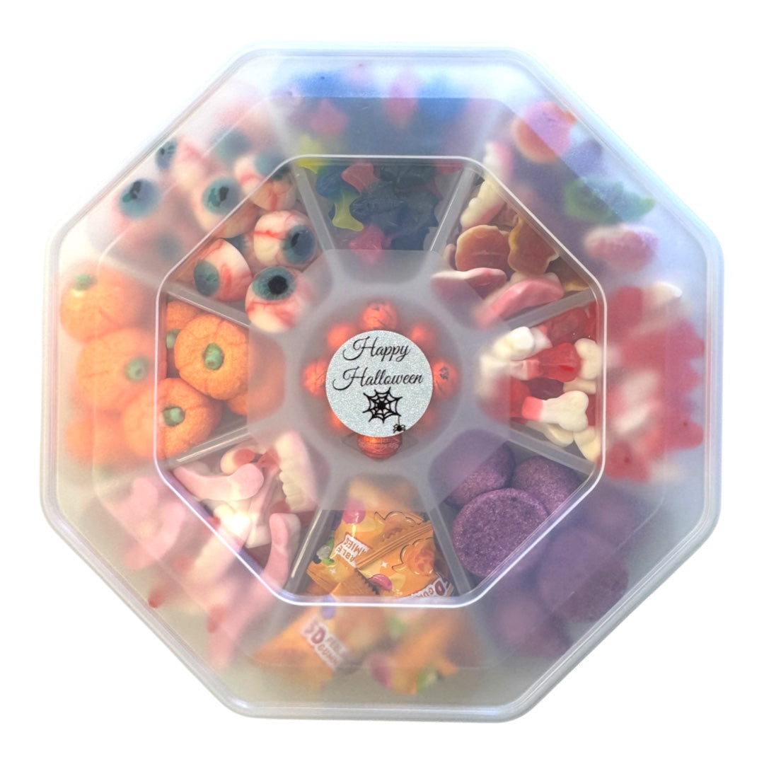 An octagonal clear plastic container filled with assorted Halloween-themed candies and gummy sweets, including orange pumpkins, purple candies, pink gummy worms, and a central round sticker that reads 'Happy Halloween'.