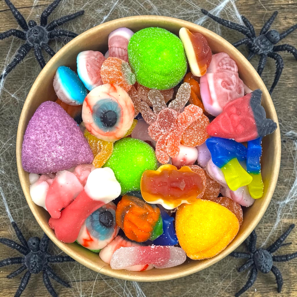 A top-down view of a bowl filled with assorted Halloween-themed gummy and hard candies, surrounded by black plastic spiders on a rustic wood surface with cobweb decorations.