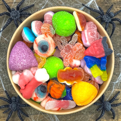 A top-down view of a bowl filled with assorted Halloween-themed gummy and hard candies, surrounded by black plastic spiders on a rustic wood surface with cobweb decorations.