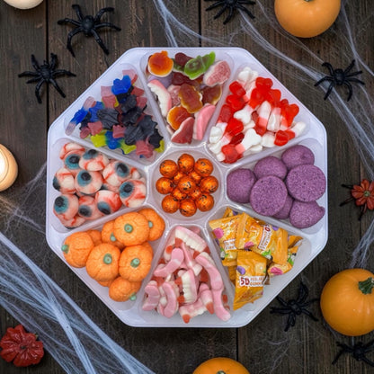 An octagonal plastic tray filled with assorted Halloween-themed candies and gummy sweets, arranged on a dark wooden surface with spider decorations, fake cobwebs, and orange pumpkins around it.