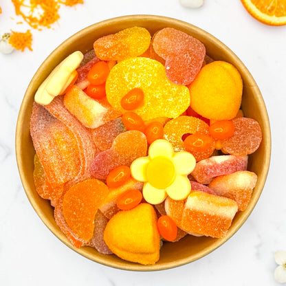 A top-down view of a candy bowl filled with assorted colourful gummy candies, including orange and yellow sweets, on a white marble surface with orange slices in the corner.