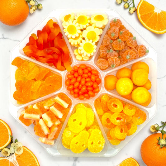 An octagonal clear plastic candy organizer filled with assorted colorful gummy candies, arranged in eight compartments on a white marble surface with orange slices and small flowers around it.