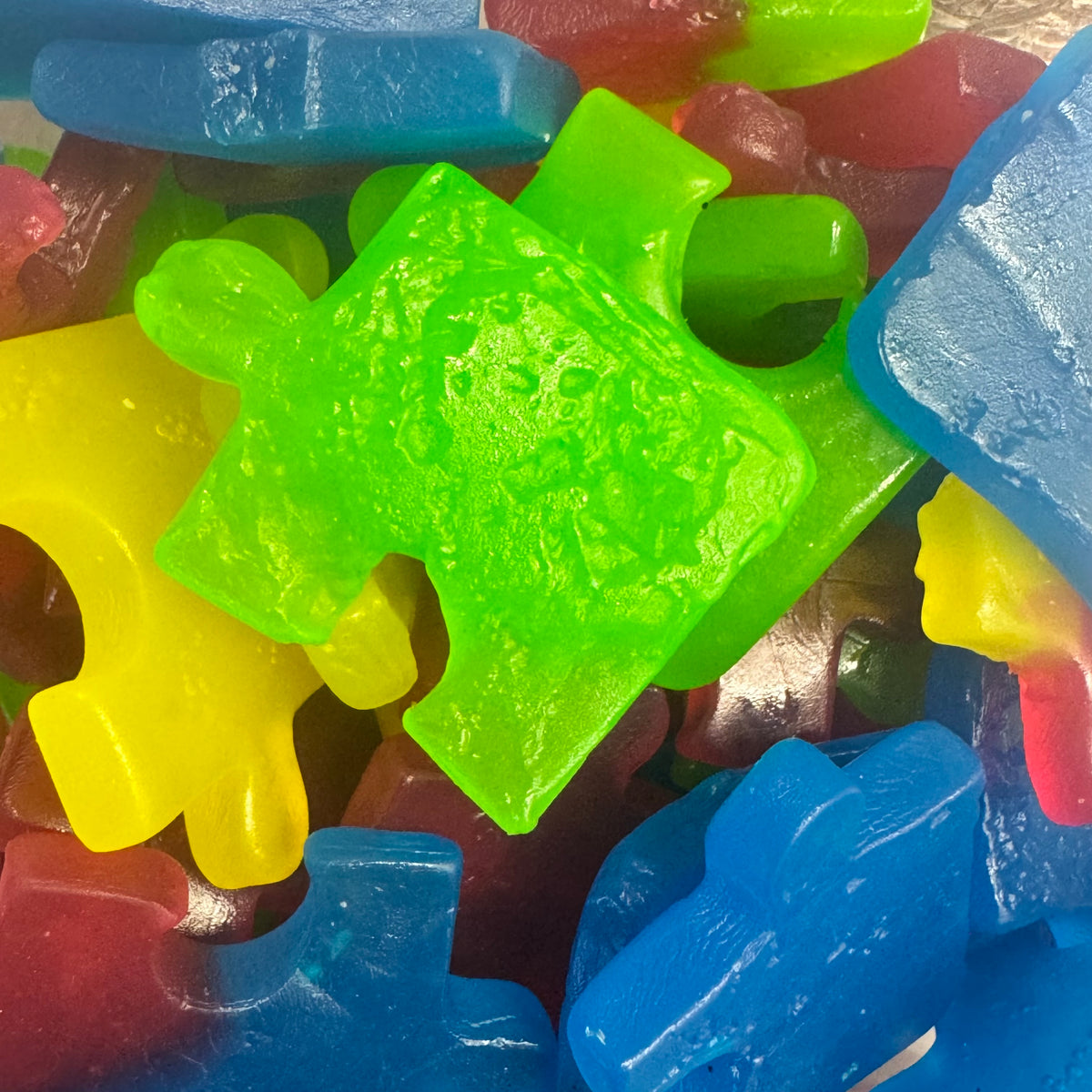 A close-up pile of translucent gummy candies shaped like jigsaw puzzle pieces, shown in bright neon green, blue, yellow, and red.