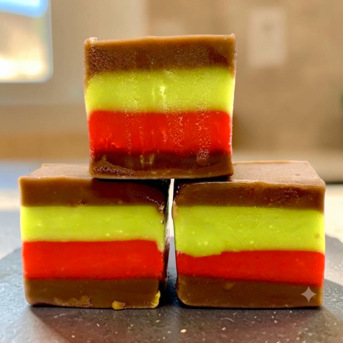Three stacked The Fudge Factory Strawberry & Pistachio Creme Sandwich Fudge, photographed on a dark surface with a softly blurred indoor background.