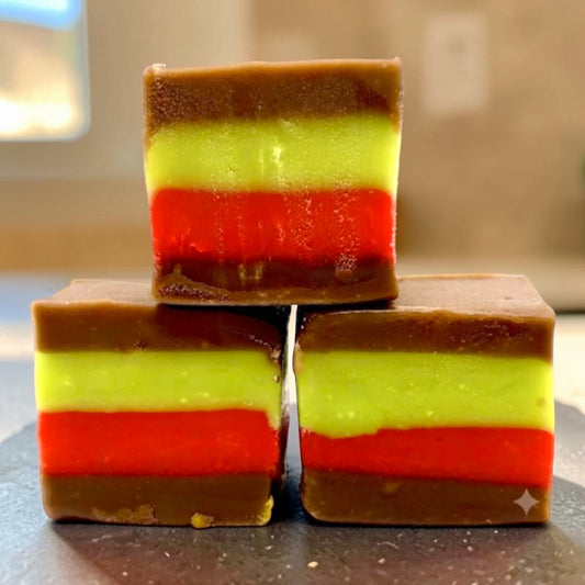 Three stacked The Fudge Factory Strawberry & Pistachio Creme Sandwich Fudge, photographed on a dark surface with a softly blurred indoor background.
