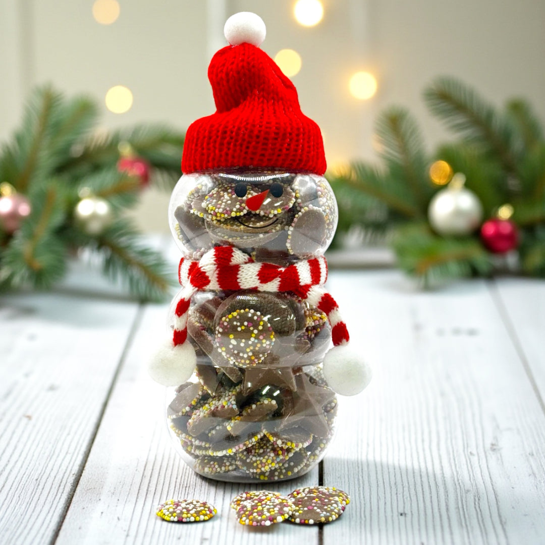 Christmas Snowman Jar filled with Milk Chocolate Jazzle's and Stars 300g. The background features a rustic table and Christmas Foliage